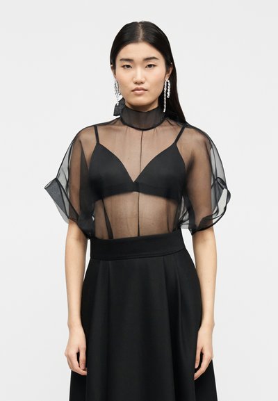 Femme aux longs cheveux noirs portant un haut noir transparent à col haut sur un soutien-gorge noir, une jupe noire et de longues boucles d'oreilles pendantes, debout devant un fond blanc.