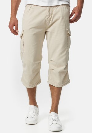 Beige Cargo-Shorts mit zwei Seitentaschen, knielangem Saum und genähten Details. Getragen mit weißen Turnschuhen.