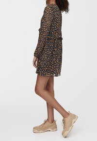 Robe fleurie bleu foncé avec des fleurs orange et jaunes, manches longues, ceinture à la taille et ourlet à volants. Portée avec des baskets beiges épaisses.