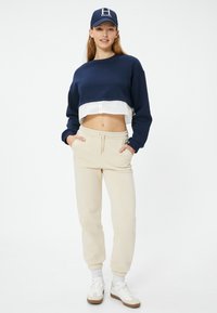 Marineblaues, kurzes Sweatshirt über einer weißen Bluse, kombiniert mit beigen, schmal zulaufenden Jogginghosen und weißen Sneakers mit braunen Akzenten.
