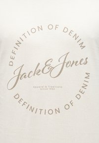 Μπεζ ύφασμα με το κείμενο «Jack & Jones» στο κέντρο, περιβαλλόμενο από κυκλικό κείμενο «Definition of Denim» και την επιγραφή «Apparel & Traditions Since 1990» από κάτω.