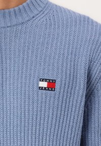 Lichtblauwe gebreide trui met een ribbelstructuur. Heeft een kleine, multicolore logo patch op de borst met de tekst "TOMMY JEANS."