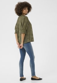 Olīvu zaļas mežģīņu blūze ar pūkainām piedurknēm, kombinēta ar pieguļošām zilām džinsām un melniem baleta apaviem. Auduma virspusē ir strukturēts ziedu raksts.