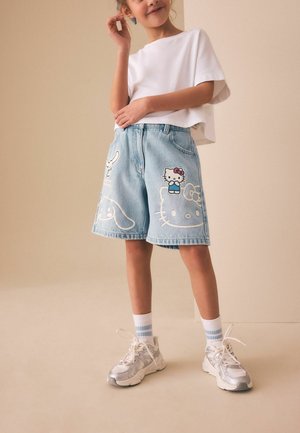 Gyermek világoskék farmer rövidnadrágban Hello Kitty mintákkal, fehér pólóban, csíkos zokniban és ezüst színű tornacipőben, bézs háttér előtt áll.