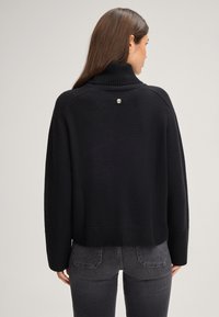 Maglione a collo alto nero con un motivo a maglia testurizzato, design cropped e un bottone d'accento argentato sul retro. Abbinato a jeans grigio scuro.