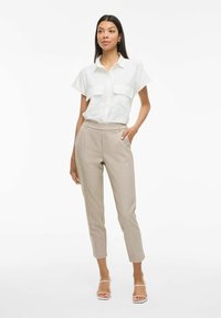 Chemise blanche à manches courtes avec poches à rabat, associée à un pantalon beige clair taillé. Le modèle porte des sandales transparentes àlanières.