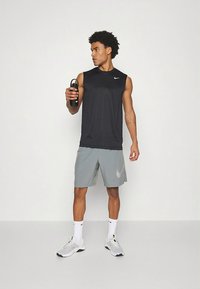 Schwarzes ärmelloses Nike-Shirt, hellgraue Shorts mit dezentem Logo, weiße Sportsocken und weiße Schuhe mit gelben Akzenten, hält eine schwarze Wasserflasche.
