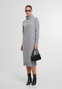 Grau gestricktes Kleid mit hohem Kragen und gerippten Bündchen, kombiniert mit einer kleinen schwarzen Handtasche und glänzenden schwarzen Stiefeletten.