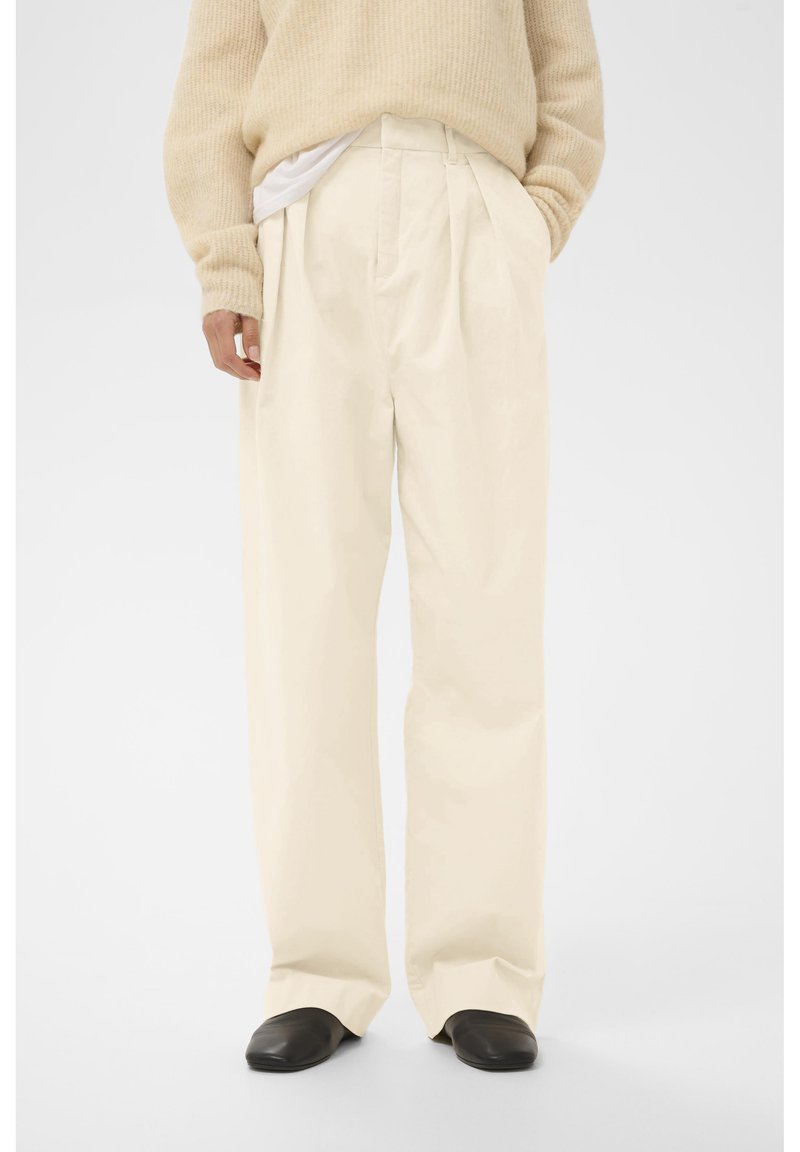 Persoană îmbrăcată cu pantaloni largi de culoare crem, pulover bej parțial băgat în pantaloni și pantofi negri, stând în fața unui fundal alb simplu.