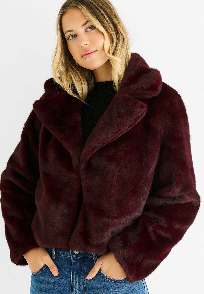 Burgundy faux fur crop jacket with a wide collar and long sleeves, paired with a black textured top and blue jeans, hands in pockets.