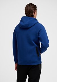 Modrá mikina s utiahnutou kapucňou, hladkým povrchom a detailom bočného zipsu. V kombinácii s čiernymi nohavicami vytvára pohodlný outfit.