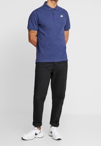 Marinblå polo med krage och en vit Nike-logga, matchad med svarta uppvikta byxor och vita sneakers med svarta accenter.