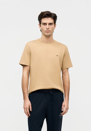 Άνδρας όρθιος, φοράει μπεζ κοντομάνικη μπλούζα με μικρό λογότυπο στο στήθος και μαύρο παντελόνι με κορδόνι και λεπτομέρεια κειμένου στο πλάι.