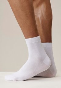 Para brązowych nóg noszących proste białe skarpetki do kostek, ustawionych z jedną stopą płasko na ziemi, a drugą lekko uniesioną na palcach.