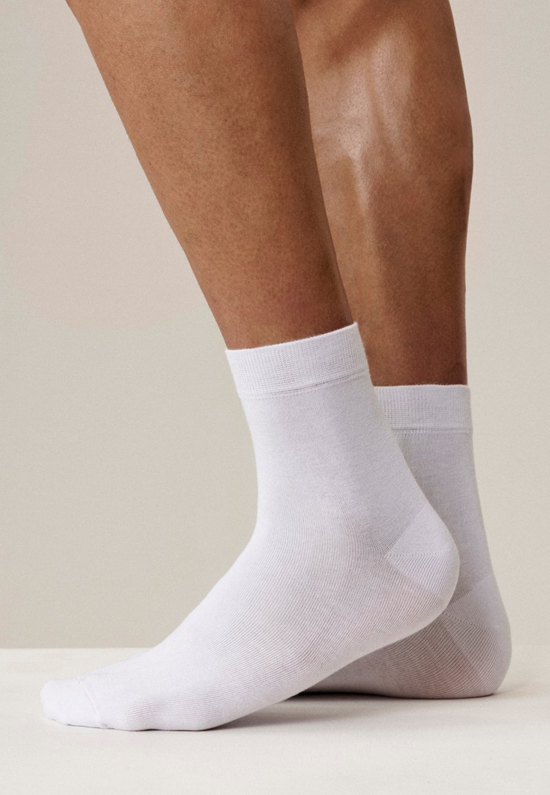 Para brązowych nóg noszących proste białe skarpetki do kostek, ustawionych z jedną stopą płasko na ziemi, a drugą lekko uniesioną na palcach.