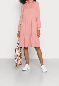 Roze lange jurk met een gerimpelde halslijn, gelaagd ontwerp en een ontspannen pasvorm. Gecombineerd met een gebloemde tas en witte sneakers.