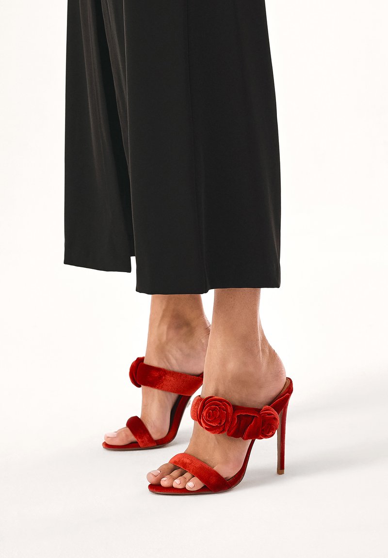 Sandales à talons hauts en velours rouge avec une large bride ornée d'une accent floral, bout ouvert et talon aiguille fin. Associe avec un pantalon noir à jambes larges.