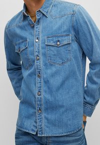 Camisa de mezclilla azul claro con dos bolsillos en el pecho, cierre de botones en la parte delantera y cuello puntiagudo. Tejido texturizado con un acabado ligeramente desgastado.