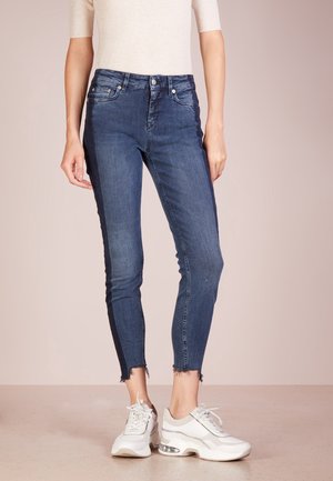 Femme portant un jean skinny bleu foncé avec des ourlets effilochés et des baskets blanches épaisses, debout devant un fond clair uni.