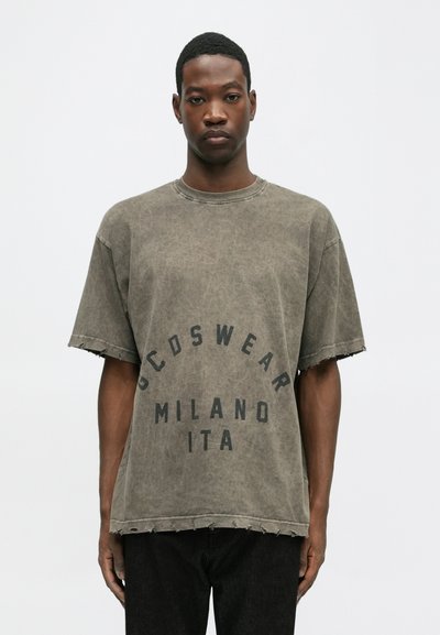 Tricou oversized din bumbac gri, cu o margine deteriorată, având text negru "GCDSWEAR MILANO ITA" imprimat într-un design circular.