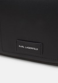 Svart lærveske med rektangulær form, utstyrt med et sydd logo-patch som leser "KARL LAGERFELD" i hvit tekst.