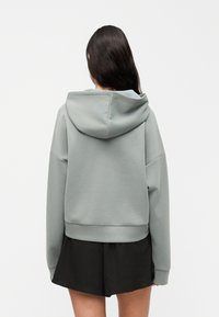 Φούτερ με κουκούλα από βαμβακερό μείγμα σε ανοιχτό πράσινο χρώμα. Χαλαρή γραμμή με χαμηλωμένους ώμους και ελαστική μέση. Συνδυασμένο με μαύρη φούστα.