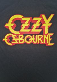 Sort stof med fed, rød og gul stiliseret tekst, der staver "OZZY OSBOURNE," med karakteristiske vinklede former og konturer.