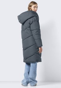 Manteau long rembourré en gris foncé avec une capuche, présentant un matelassage en chevron et des manches longues, associé à un jean large bleu clair.
