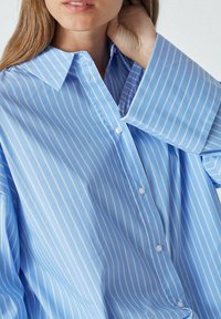 Light blue button-up shirt featuring white vertical stripes, a pointed collar, and wide sleeves. Made of smooth fabric with button accents.