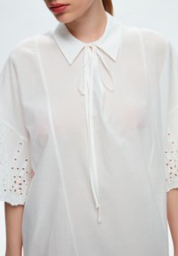 Chemise blanche avec col pointu et détail de cravate, dotée de manches courtes en dentelle texturée et d'un tissu léger.