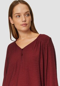 Blouse bordeaux avec un décolleté en V, présentant des détails froncés aux épaules et deux boutons décoratifs au niveau du col. Tissu texturé.