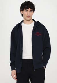 Marinblå zip-up hoodie i mjukt tyg med kängurufickor, ribbade ärmslut och broderad röd text på vänster bröst. Bärs över en vit t-shirt.