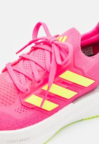 Chaussure de sport rose vif avec des bandes jaunes, empeigne tricotée, lacets attachés et semelle blanche avec un détail d’amorti léger Boost.