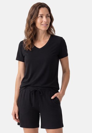 Donna con capelli castani mossi che indossa una maglietta nera a scollo a V e pantaloncini neri con coulisse, in piedi con una mano in tasca, guarda a destra.