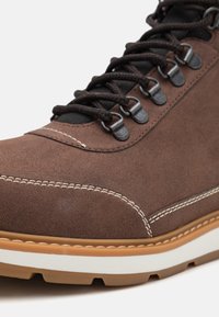 Bota de cuero sintético marrón con costuras blancas, ojales de metal y una superficie texturizada. Suela de goma con acentos en tono marrón claro.