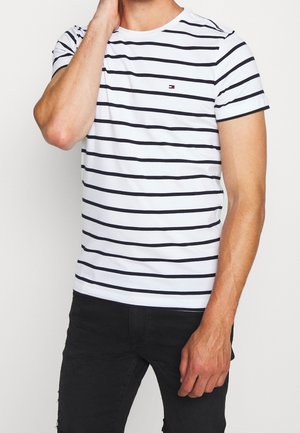 Man die een wit T-shirt met korte mouwen draagt met zwarte horizontale strepen en een klein logo, gecombineerd met een zwarte broek.