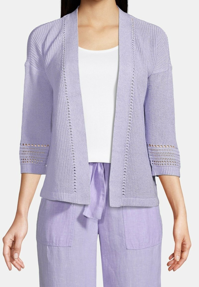 Femme portant un cardigan tricoté violet clair ouvert sur le devant avec des détails en œillets sur les manches, sur un haut blanc et un pantalon violet assorti.