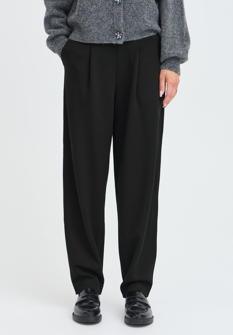 Personne portant un pantalon noir large, des chaussures noires et un pull gris boutonné avec les mains partiellement dans les poches.