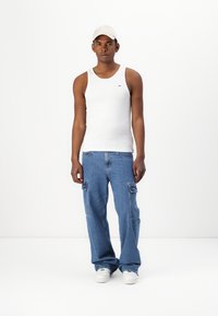 Hvit ermeløs tank top, blå cargo jeans med lommer, og hvite joggesko. Modell som har på seg en lysfarget caps mot en ensfarget bakgrunn.