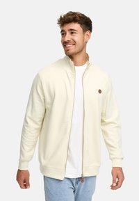 Hombre sonriente con cabello corto y rizado, vistiendo una chaqueta cremosa con cremallera sobre una camisa blanca y jeans azul claro, mirando hacia la derecha.