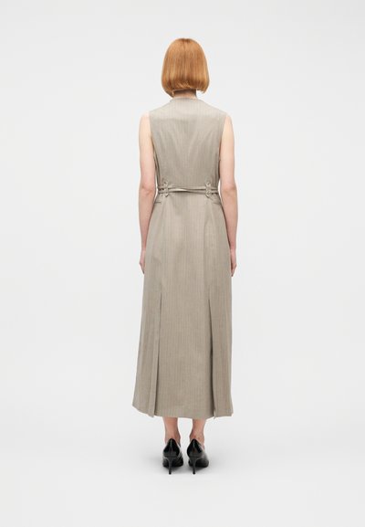 Γυναίκα με κοντά κόκκινα μαλλιά που φοράει μπεζ midi φόρεμα με ριγέ μοτίβο χωρίς μανίκια και μαύρα ψηλοτάκουνα, που στέκεται γυρισμένη προς τα πίσω μπροστά σε λευκό φόντο.