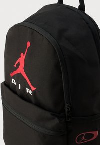 Μαύρο σακίδιο πλάτης κατασκευασμένο από ανθεκτικό ύφασμα, με κόκκινο λογότυπο Jumpman, κείμενο "AIR"  και μπροστινή τσέπη με φερμουάρ και μαύρα εξαρτήματα.