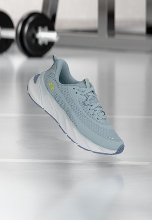 Chaussure de sport bleu clair avec semelle blanche épaisse et logo jaune, montrée flottant à l'intérieur sur un sol réfléchissant près d'équipements de gym.