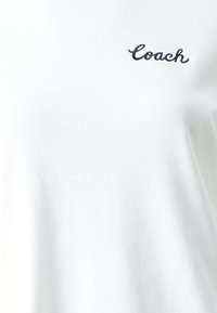 White cotton t-shirt featuring a small black embroidered "Coach" logo on the upper left side. Soft texture and standard fit design.