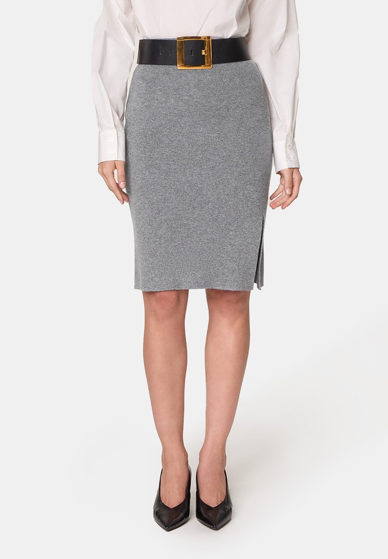 Grauer Strick-Bleistiftrock mit einem seitlichen Schlitz, kombiniert mit einem breiten schwarzen Gürtel mit einer goldenen quadratischen Schnalle. Dazu werden spitze schwarze High Heels getragen.