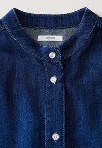 Camicia di jeans di colore blu scuro, con scollo rotondo, chiusura a bottoni con bottoni bianchi e cuciture a contrasto lungo i bordi.