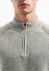 Grijze, ribbelige pullovertrui met een hoge kraag en korte rits. Textuurstof met contrasterende ribbelaccenten bij de schouders.