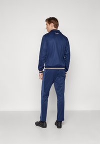 Marine trainingsjack met gestreepte manchetten en bijpassende broek; zachte stof, elastische tailleband en zichtbare schoenaccenten.
