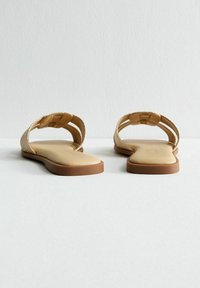 Sandales plates avec un design de lanière tissée beige, semelle intérieure lisse couleur noisette et semelle extérieure brun clair ; ouverture à l'arrière pour un enfilage facile.