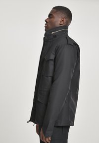 Black, multi-pocketed jacket with a high collar, featuring a zipper and button hardware. Smooth texture and streamlined design.